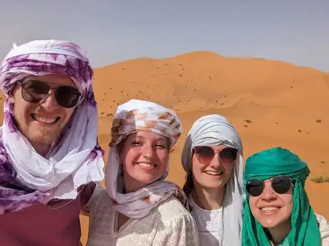 Group of travelers wearing traditional turbans smiling in front of Sahara dunes on a Sahara Desert 8 days tour from Marrakech