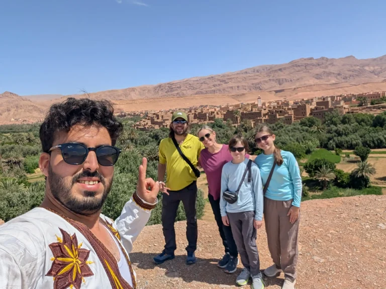Is Morocco safe for families with children? A family of four with their teenage kids and private Moroccan driver-guide pose for a selfie overlooking a palm oasis and Berber kasbah.