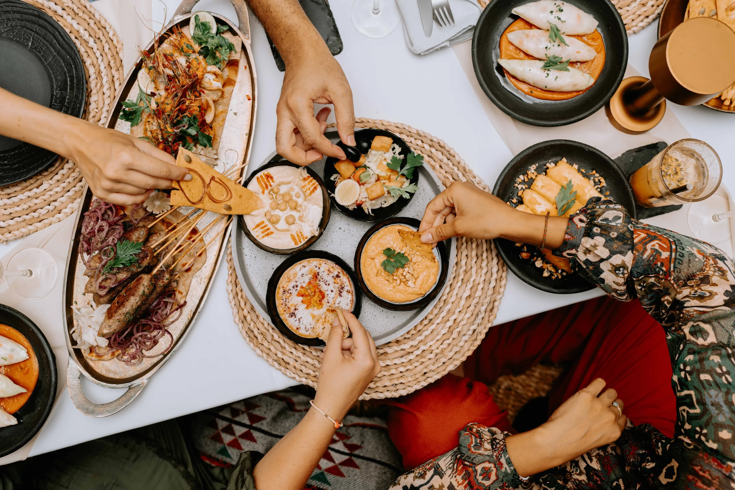 Overhead view of people sharing a Moroccan food and Mediterranean feast with dips, grilled skewers, pita bread, and seafood